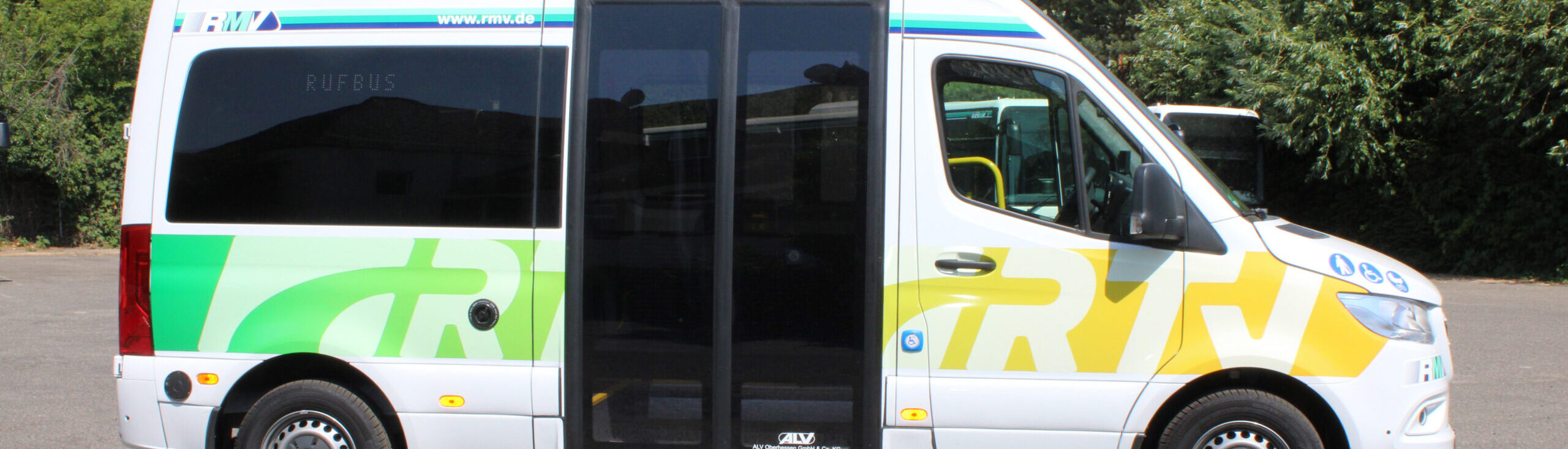 Seitliche Gesamtansicht eines modernen Rufbusses mit großem Schiebetürbereich, gelb-grüner RTV-Grafik und RMV-Logo. Auf der Tür sind Piktogramme für Barrierefreiheit, Kinderwagen und Fahrradmitnahme zu sehen.